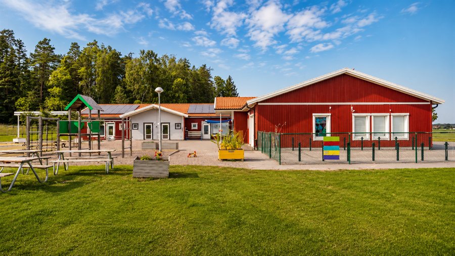 Röd och vit förskolebyggnad med lekställning, planteringar och gräsyta under blå himmel.