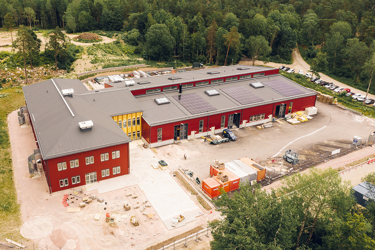Flygbild över faluröda skolbyggnader med grå tak under uppförande, omgivna av skog och landsbygd.