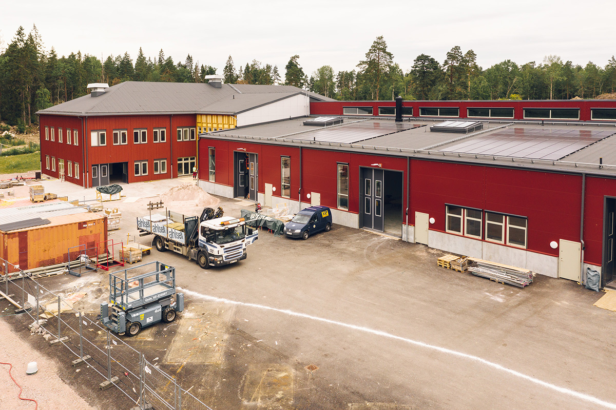 Skolgård under byggskede med faluröda byggnadskroppar, solcellstak och arbetsfordon.