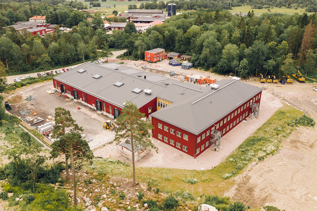 Flygbild över Jällas faluröda skolbyggnad under uppförande, omgiven av skog och landsbygd.