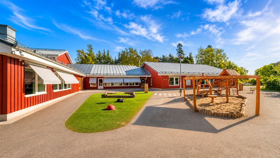 Röda förskolebyggnader kring en gård med lekställning, sandlåda och grönytor.