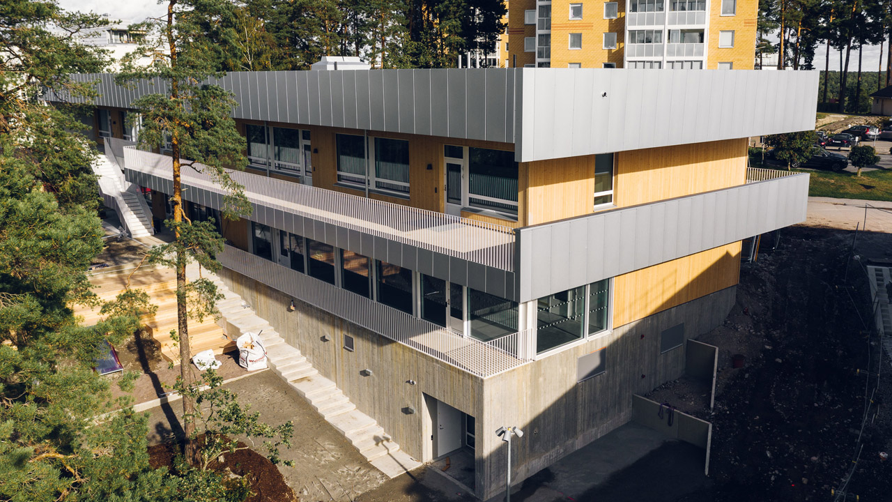 Förskolebyggnad i två plan med träfasad, plåtdetaljer och betongsockel i suterräng, med flerbostadshus i bakgrunden.